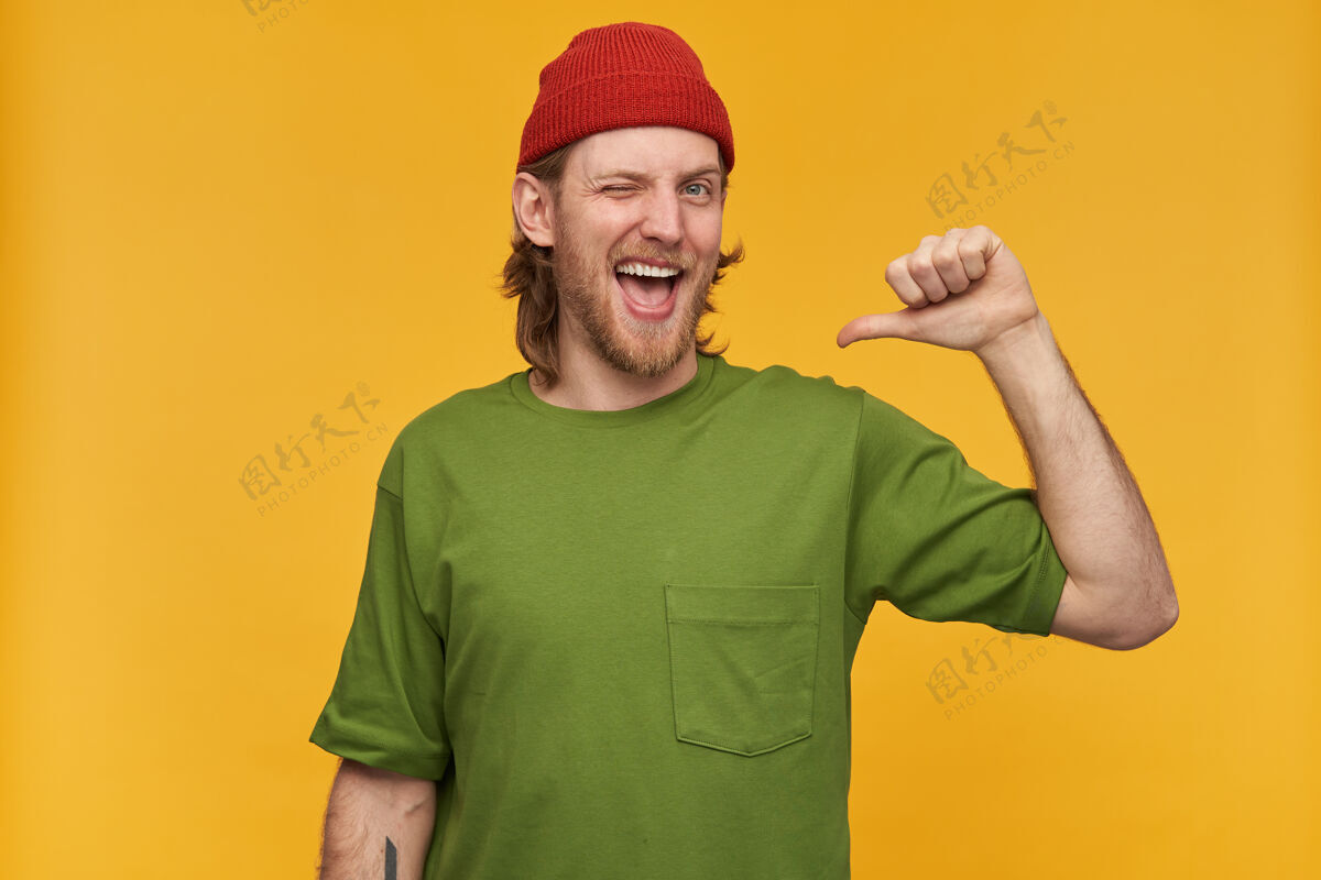 室内一幅英俊 快乐的男性肖像 金发 胡须穿着绿色t恤和红色小豆豆有纹身用拇指指着自己被隔离在黄色的墙上装束金发胡须