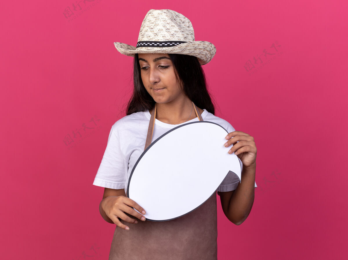 演讲穿着围裙 戴着夏帽的年轻园丁女孩举着空白的演讲泡泡牌 严肃的脸站在粉色的墙上 一边看着年轻空白靠边站
