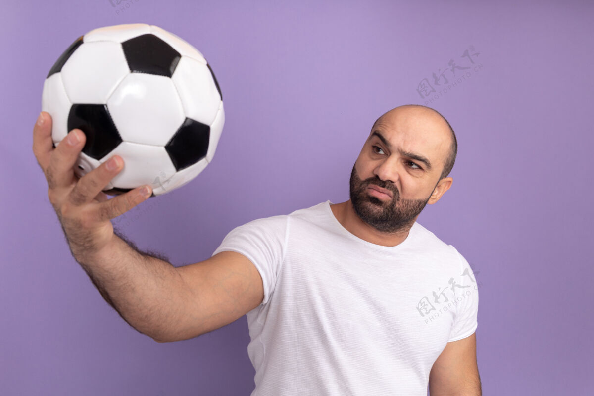 球身穿白色t恤的留胡子男人拿着足球站在紫色的墙上 带着怀疑的表情看着球怀疑足球表情