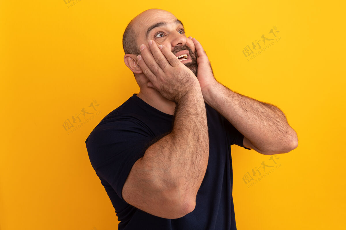 手势一个留着大胡子的穿海军蓝t恤的男人站在橘色的墙上惊讶地抬起头 双手放在脸上表情胡须男人