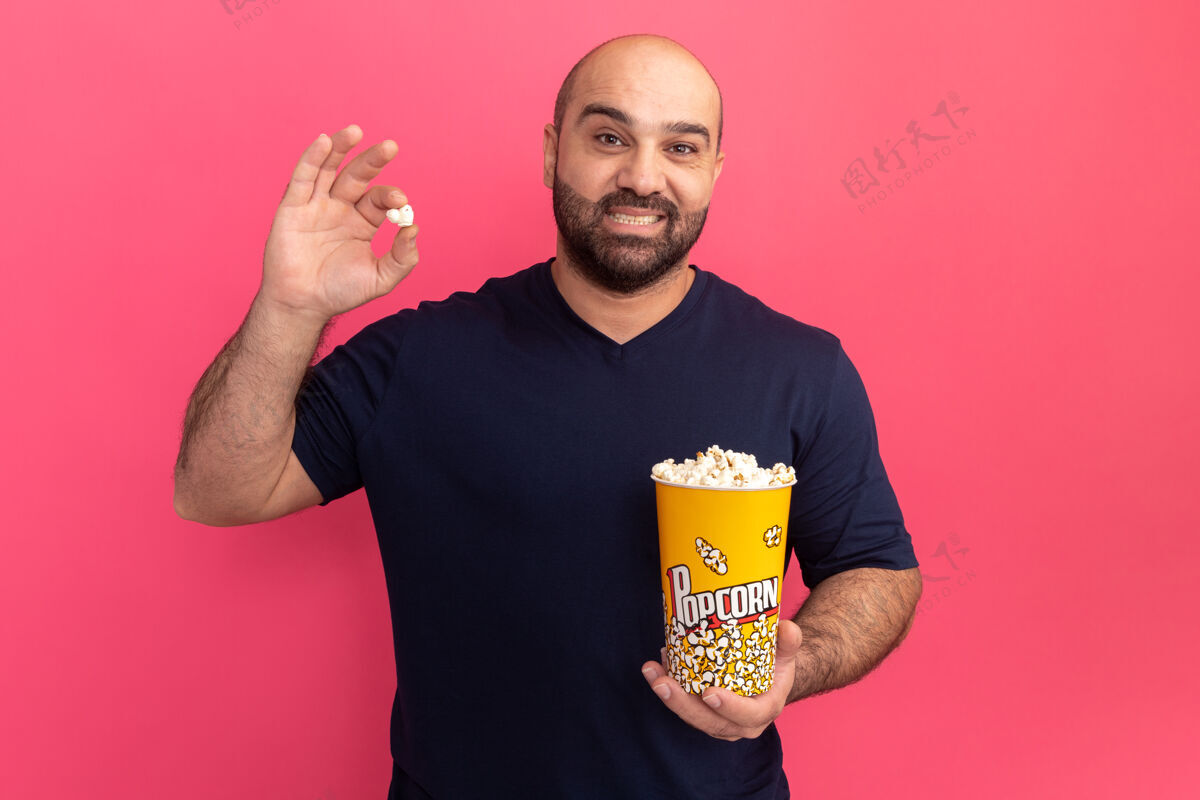 表情一个留着胡子的男人站在粉红色的墙上 穿着海军t恤 手里拿着爆米花 脸上带着微笑爆米花桶微笑