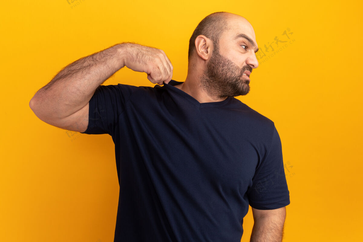 触摸一个留着大胡子的穿海军t恤的男人站在橙色的墙上 一边看着一边 一边生气地摸着他的t恤男人恼怒恼怒