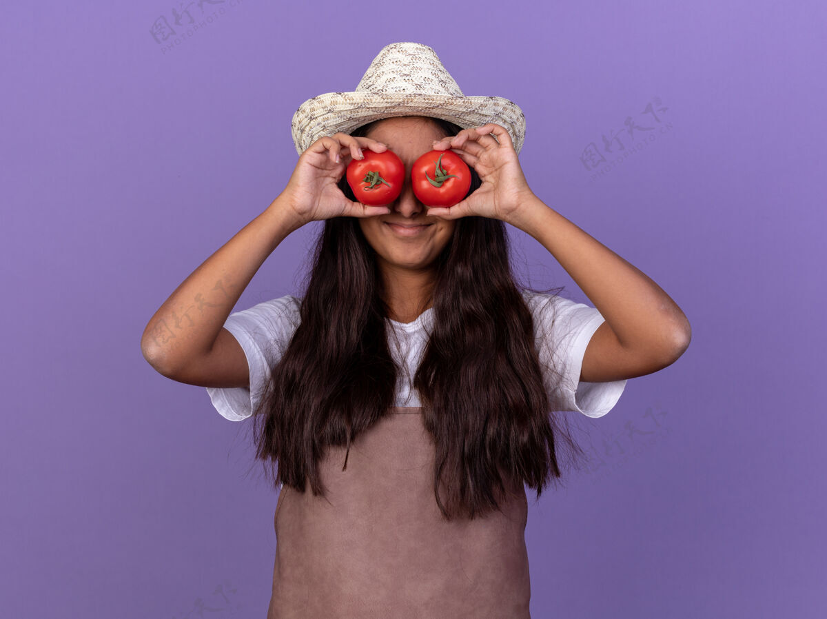 新鲜的穿着围裙 戴着夏帽 手里拿着新鲜西红柿 眼睛蒙着的年轻园丁女孩站在紫色的墙上 脸上洋溢着幸福的笑容立场围裙番茄