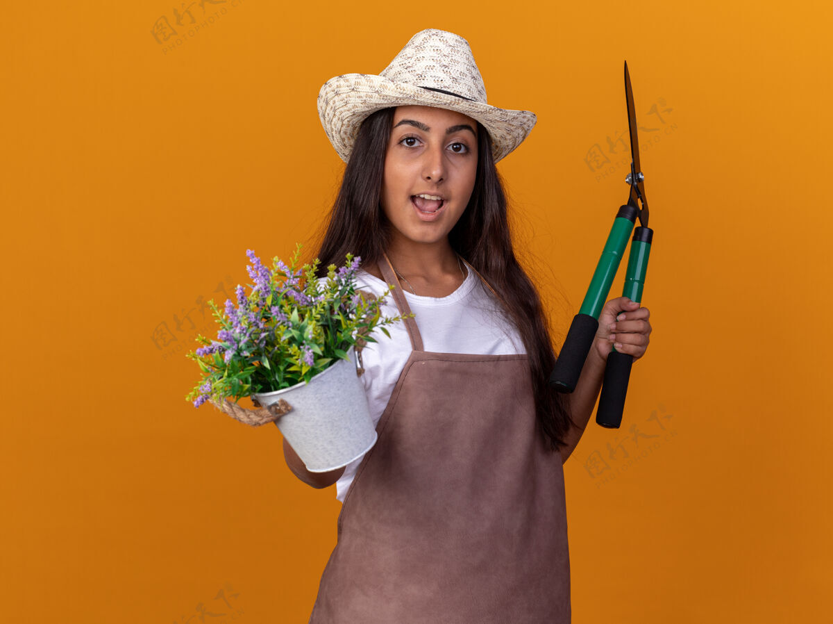 围裙穿着围裙 戴着夏帽 手持树篱剪和盆栽的年轻园丁女孩站在橙色的墙上 脸上带着微笑帽子树篱壶