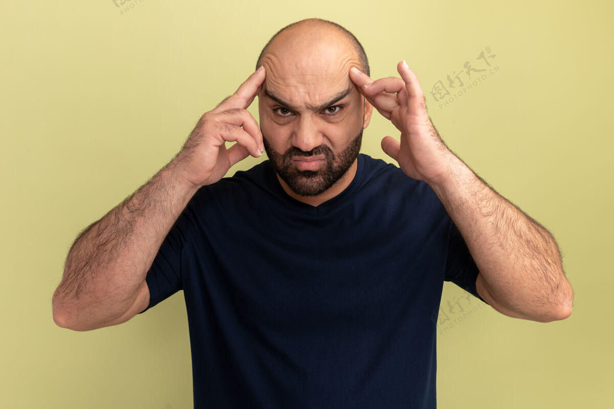 胡须一个留着胡子的男人 穿着黑色t恤 站在绿色的墙上 看着身体不舒服 摸着头很恼火 头痛得厉害手势表达头
