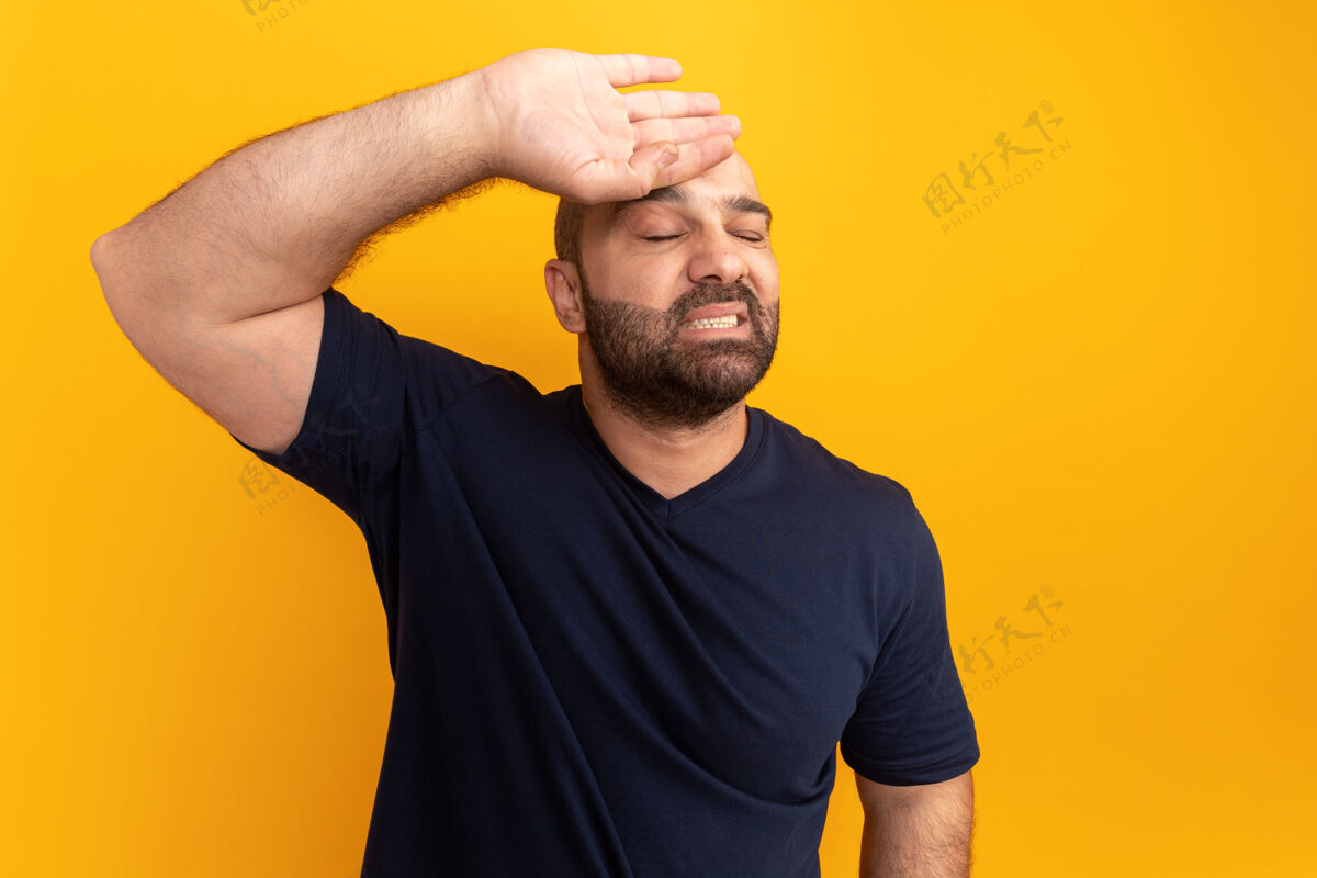 胡须一个留着胡子的男人 穿着海军蓝t恤 手放在额头上 站在橙色的墙上 脸上露出恼怒的表情表情手势男人