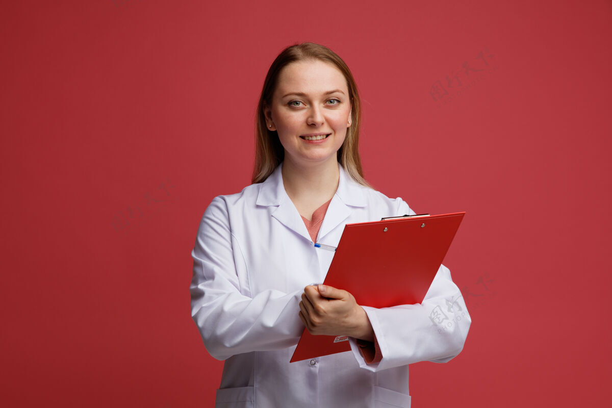 医生微笑着的年轻金发女医生穿着医用长袍 脖子上戴着听诊器 用笔在剪贴板上写字长袍金发听诊器