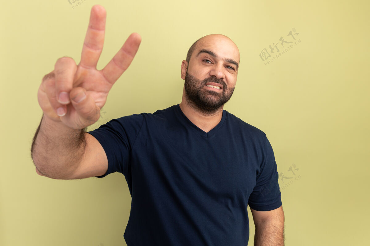 胡须快乐的留着胡子的男人穿着黑色t恤 微笑着自信地展示着站在绿色墙壁上的v形标志表演男人手势