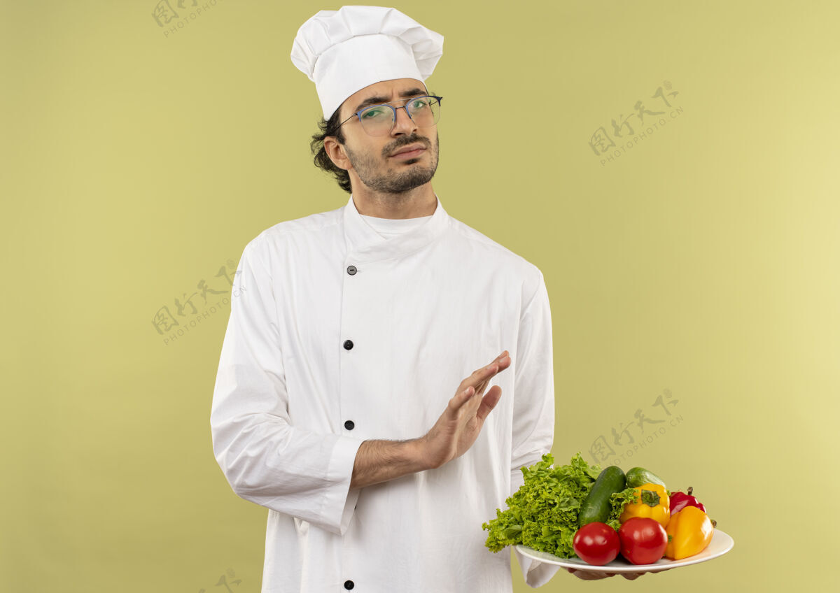 烹饪年轻的男厨师穿着厨师制服 戴着眼镜 手里拿着盘子里的蔬菜 摆着停车的姿势不愉快盘子厨师