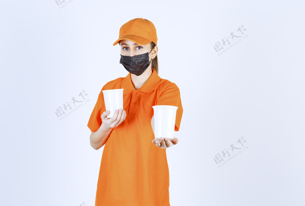 促销身穿黄色制服 戴黑色口罩的女快递员双手捧着塑料杯子里的外卖饮料 把饮料递给顾客卫生送货服装