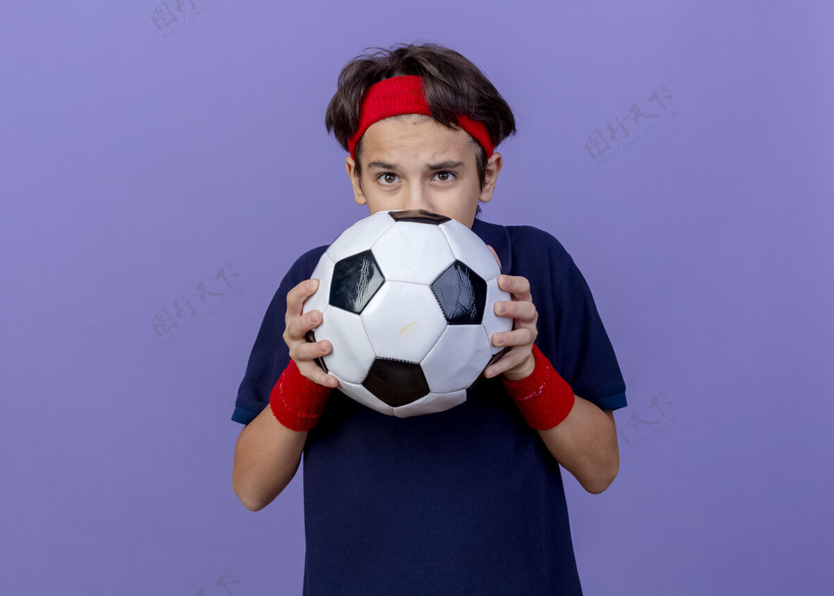 帅气令人印象深刻的年轻英俊的运动男孩戴着头带和护腕与牙套举行足球从后面它隔离在紫色墙上复制空间男孩戴运动