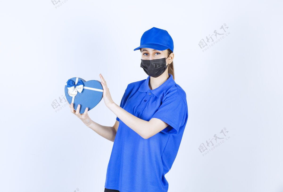 成人戴着面具和蓝色制服的女快递员手里拿着一个心形礼盒船运年轻人姿势