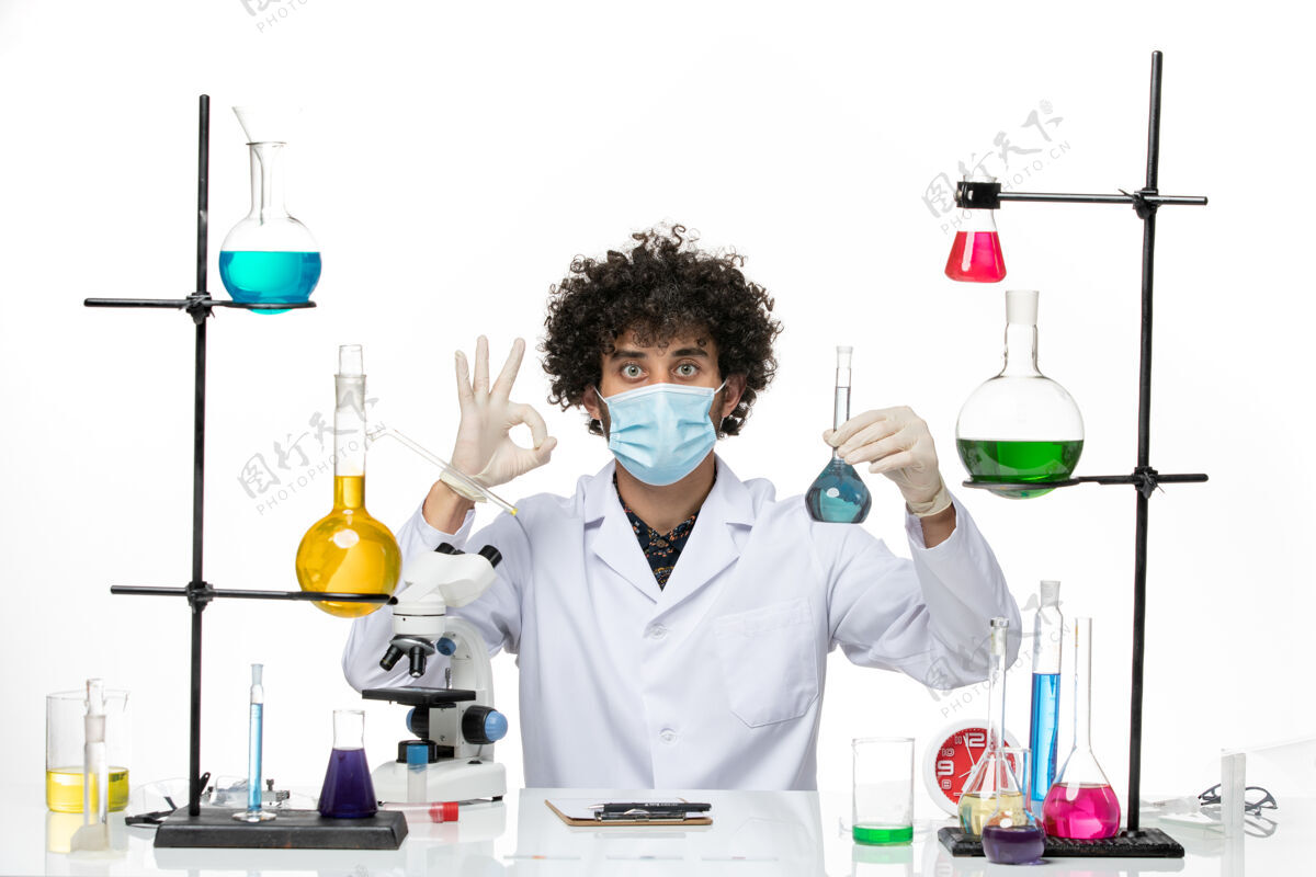 实验室外套前视图男性化学家 穿着白色医疗服 戴着面罩 在浅白的空间里拿着蓝色溶液专业医学面具