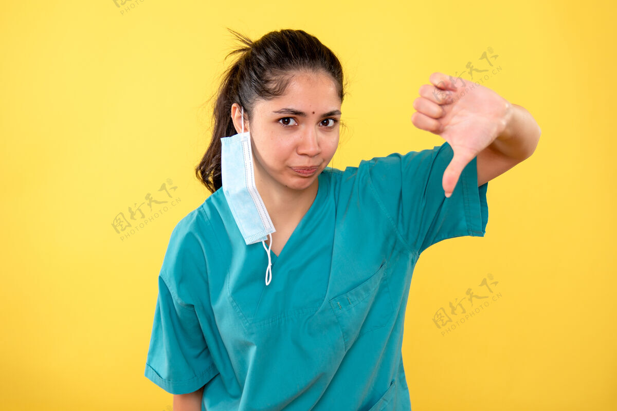 听诊器戴着医用口罩的年轻女性正面图 黄色墙上有拇指朝下的标志制造前面拇指