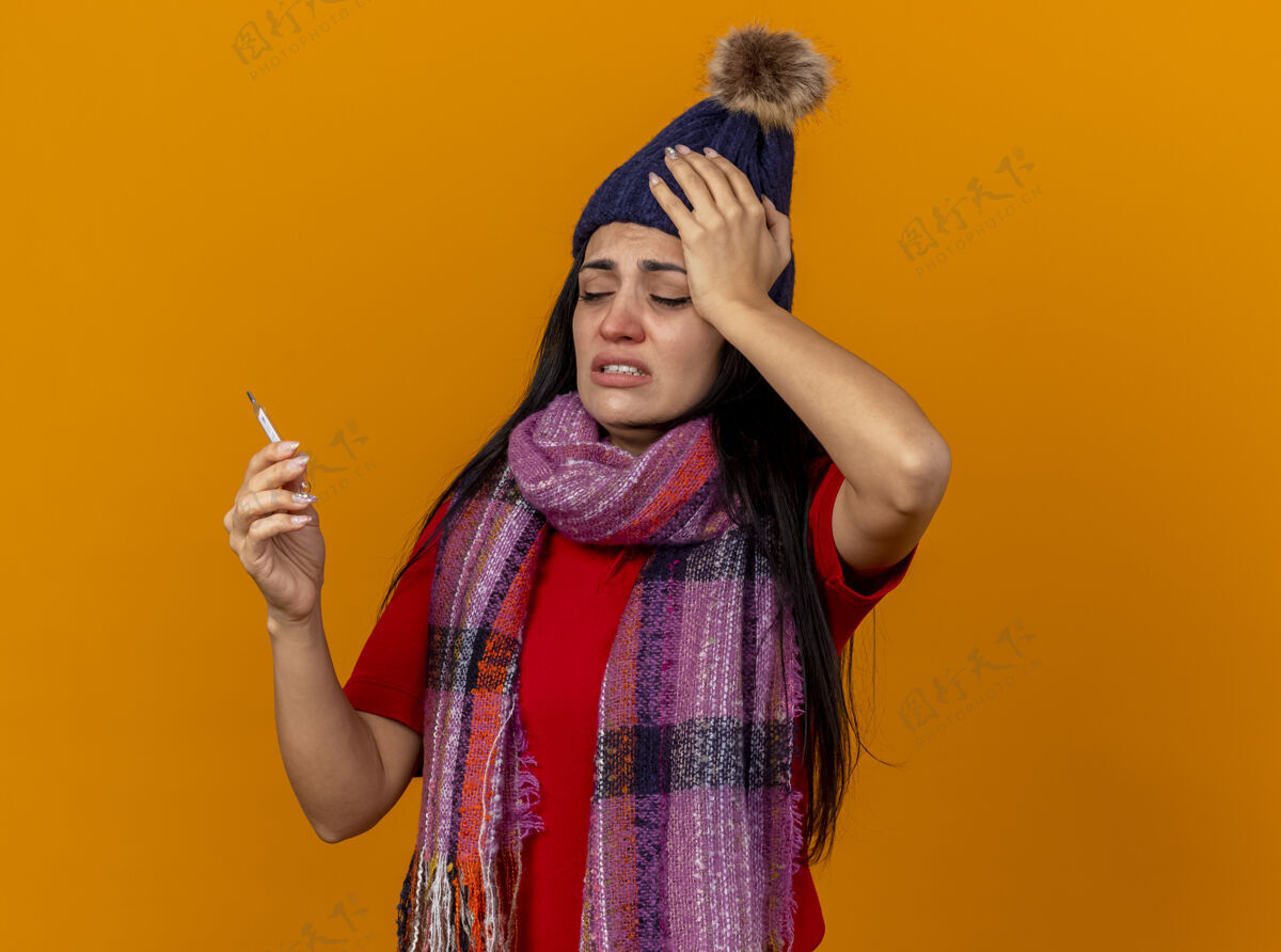 疼痛疼痛的年轻白种人生病的女孩戴着冬天的帽子和围巾拿着和看温度计隔离在橙色墙上的复制空间穿着体温计年轻