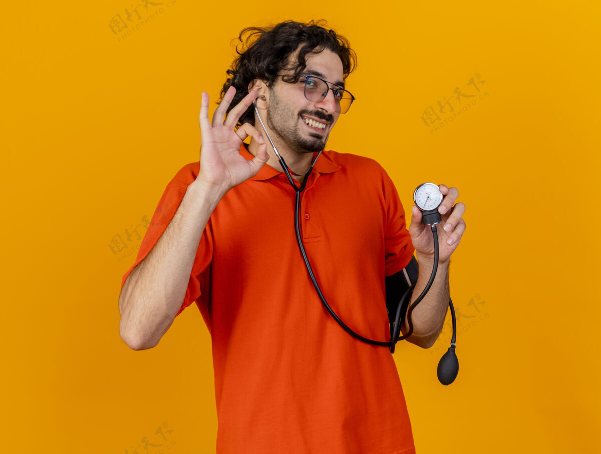 年轻带着微笑的年轻白人病人戴着眼镜和听诊器拿着血压计 在橙色的墙上隔离着“做得好”的标志听诊器橙色男人
