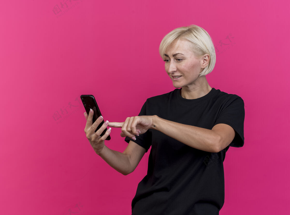 手机高兴的中年金发斯拉夫女子拿着手机看着用它隔离在深红色墙上的复制空间电话女人金发