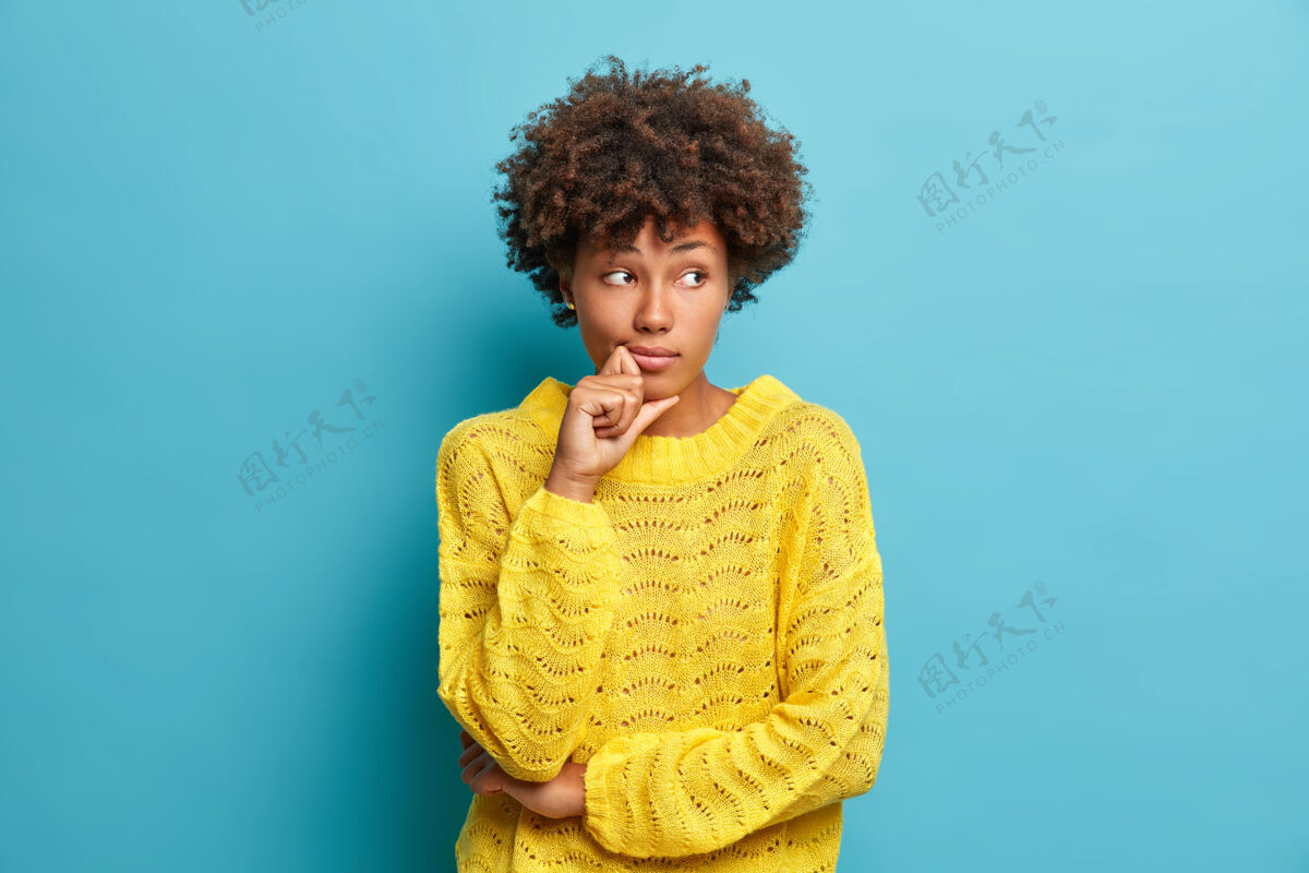 表情严肃沉思的非洲头发女人看着别处 站在沉思的姿势思考问题和困难穿着温暖的黄色毛衣隔离在蓝色的墙上做决定人思考卷发