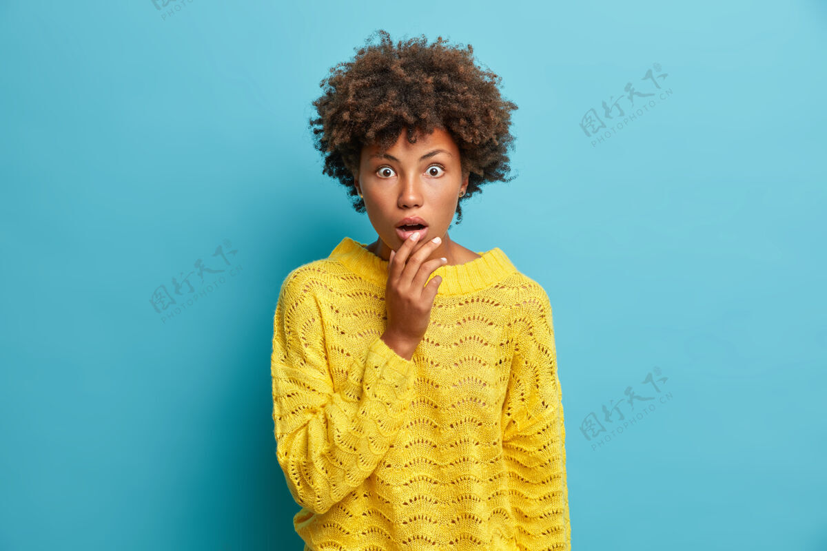 毛衣可爱惊艳的卷发女人发现惊悚新闻瞪大嘴巴站在镜头前惊悚的穿着休闲黄色套头衫隔离在蓝色的墙上发疯黑发兴奋