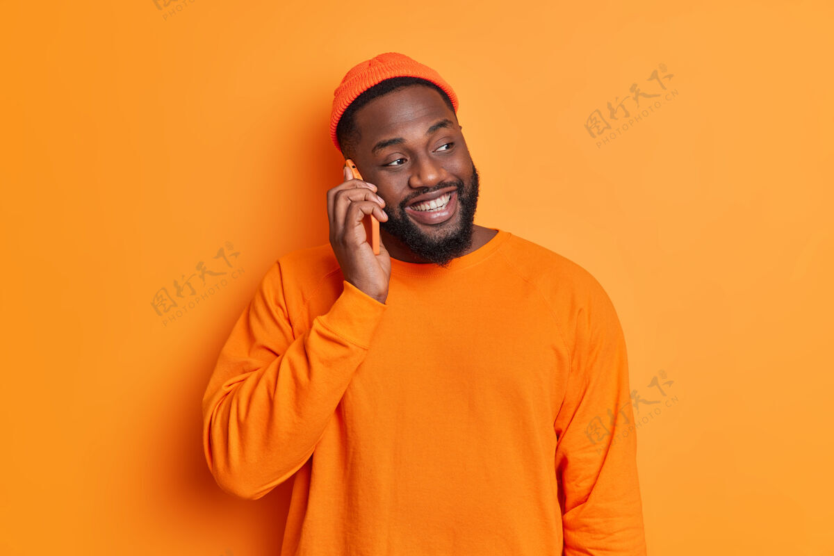 欢呼快乐胡子男人的单独镜头通过手机有愉快的谈话集中在一边微笑快乐地戴着帽子和毛衣对着橙色的工作室墙摆姿势休闲牙牙学语大笑