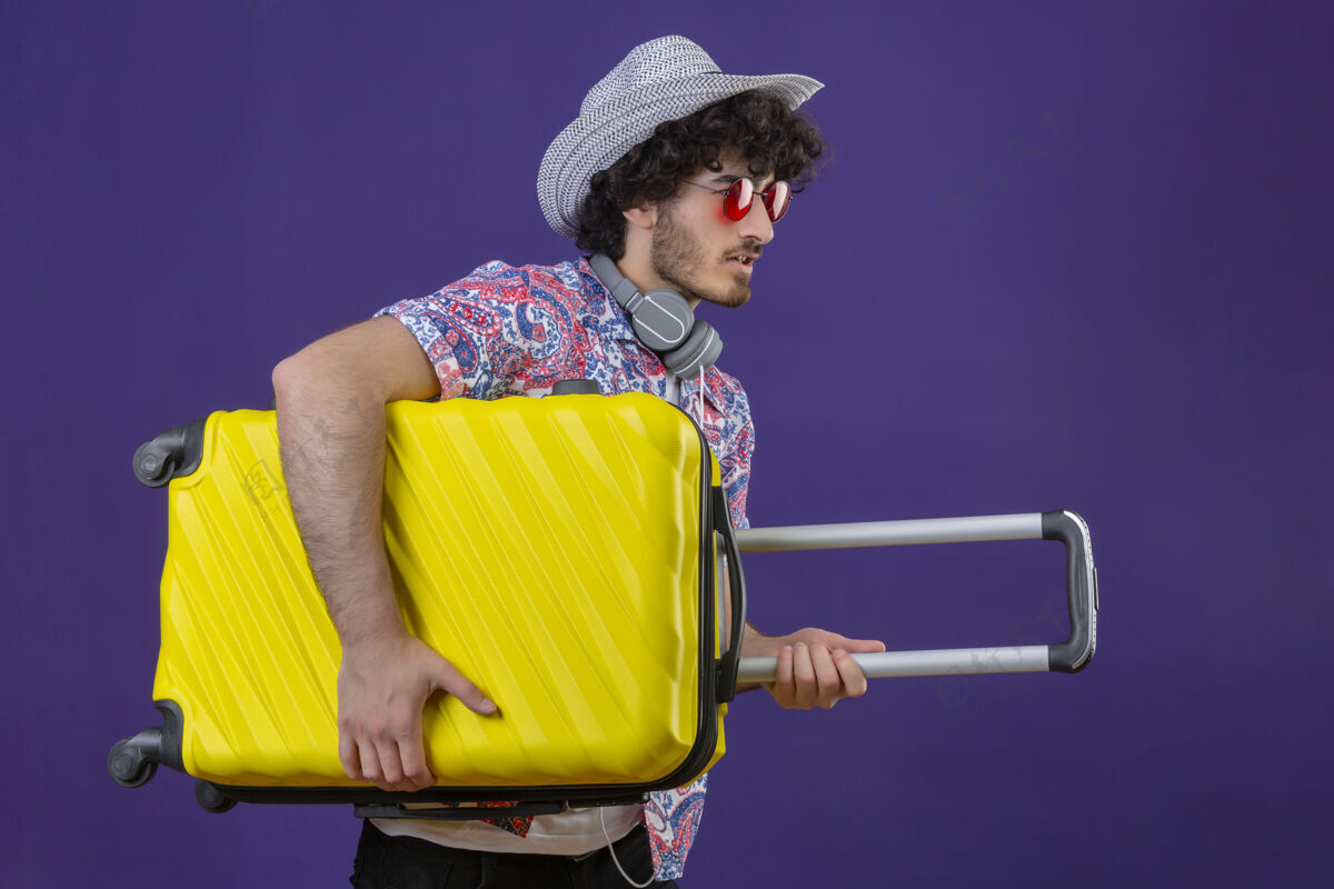 年轻年轻英俊的卷发旅行者戴着太阳镜 戴着耳机 戴着帽子 提着手提箱站在孤立的紫色墙上的侧视图上帅气立场卷发