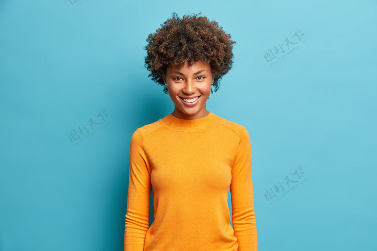 乐观半身镜头中 一位年轻而开朗的女士穿着休闲的橙色套头衫 脸上洋溢着愉快的笑容 站在蓝色的墙上有趣健康爱