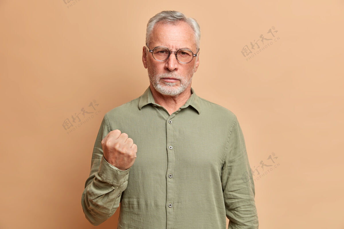 侵略性愤怒的胡须成熟男子握紧拳头 展示愤怒的画像警告你穿着正式衬衫对着米色墙壁摆姿势皱纹攻击疯狂
