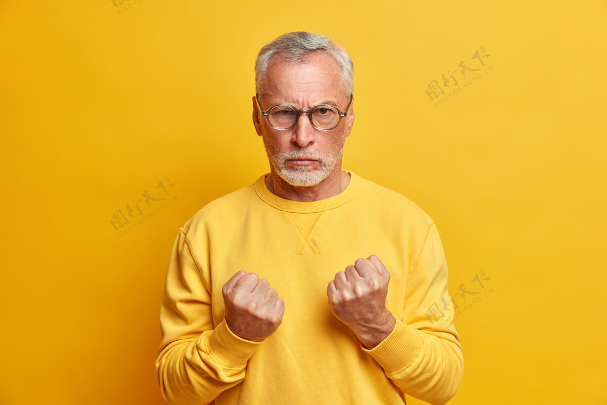 严重愤怒的老人握紧拳头 准备自卫 怒气冲冲 满脸愤慨地站在前面 穿着随便 对着黄墙摆姿势压力男人拳打