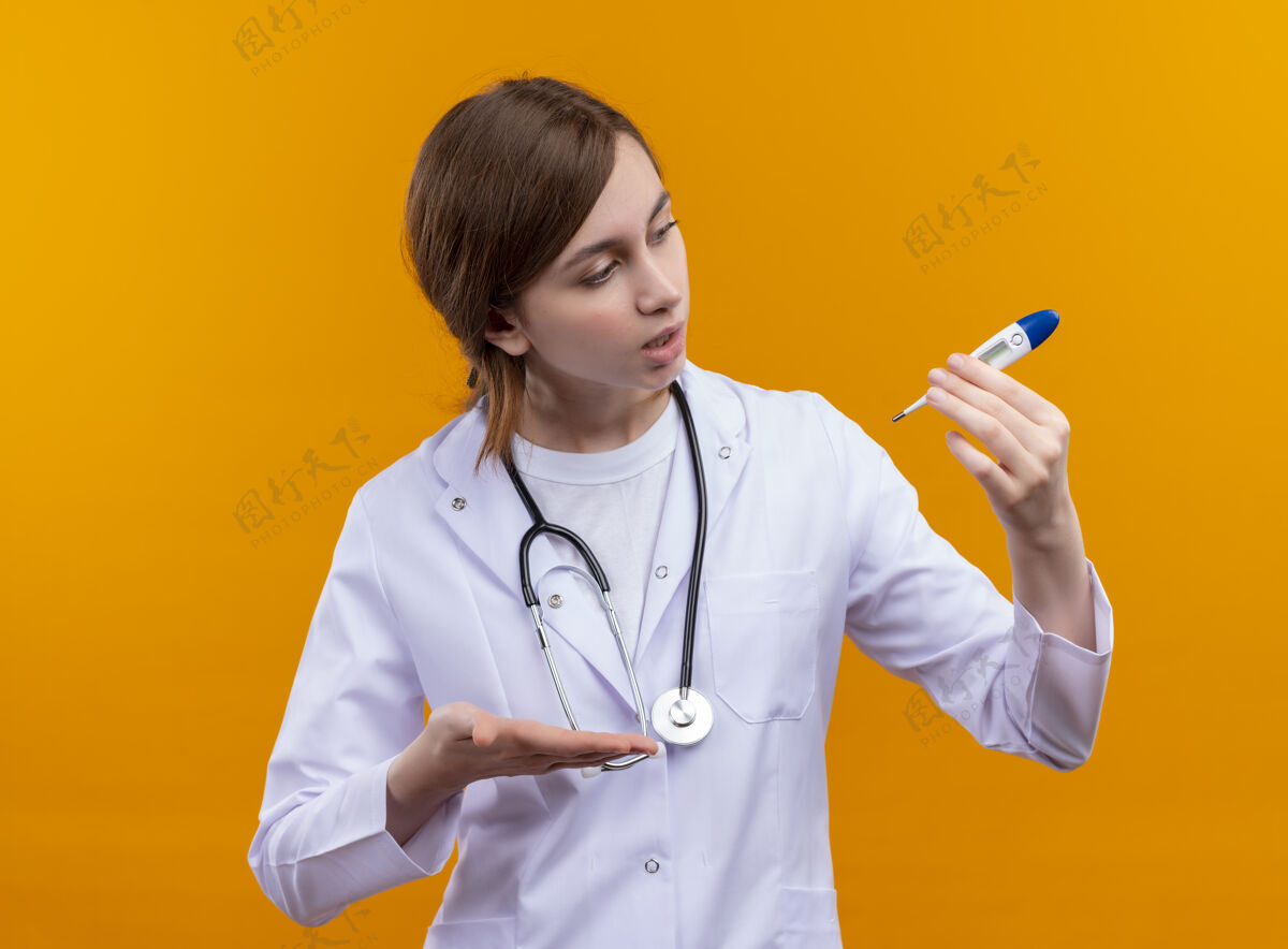 体温计年轻的女医生穿着医用长袍 手持听诊器 拿着体温计 惊讶地看着隔离的橙色墙上的温度计女听诊器穿