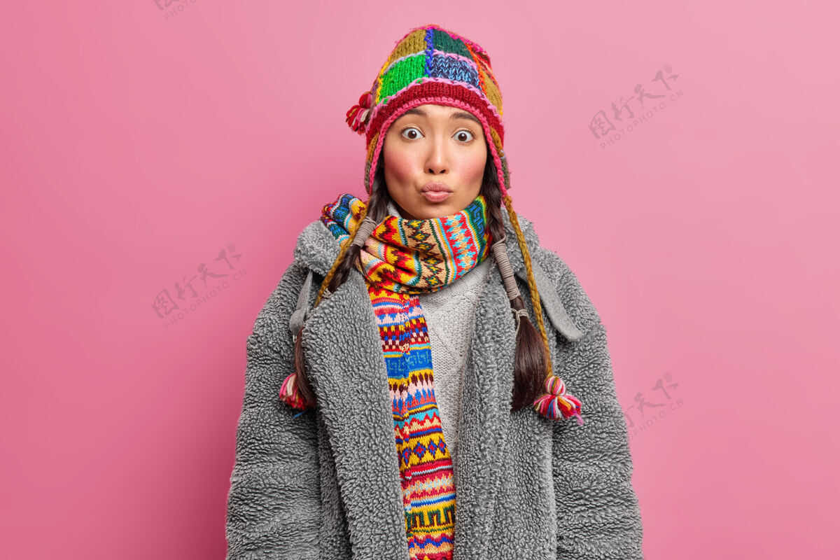 黑发年轻的亚洲女人嘴唇圆润有着惊讶的表情 戴着针织帽子和围巾 穿着灰色皮毛外套 隔着粉色的墙壁姿势惊喜衣服