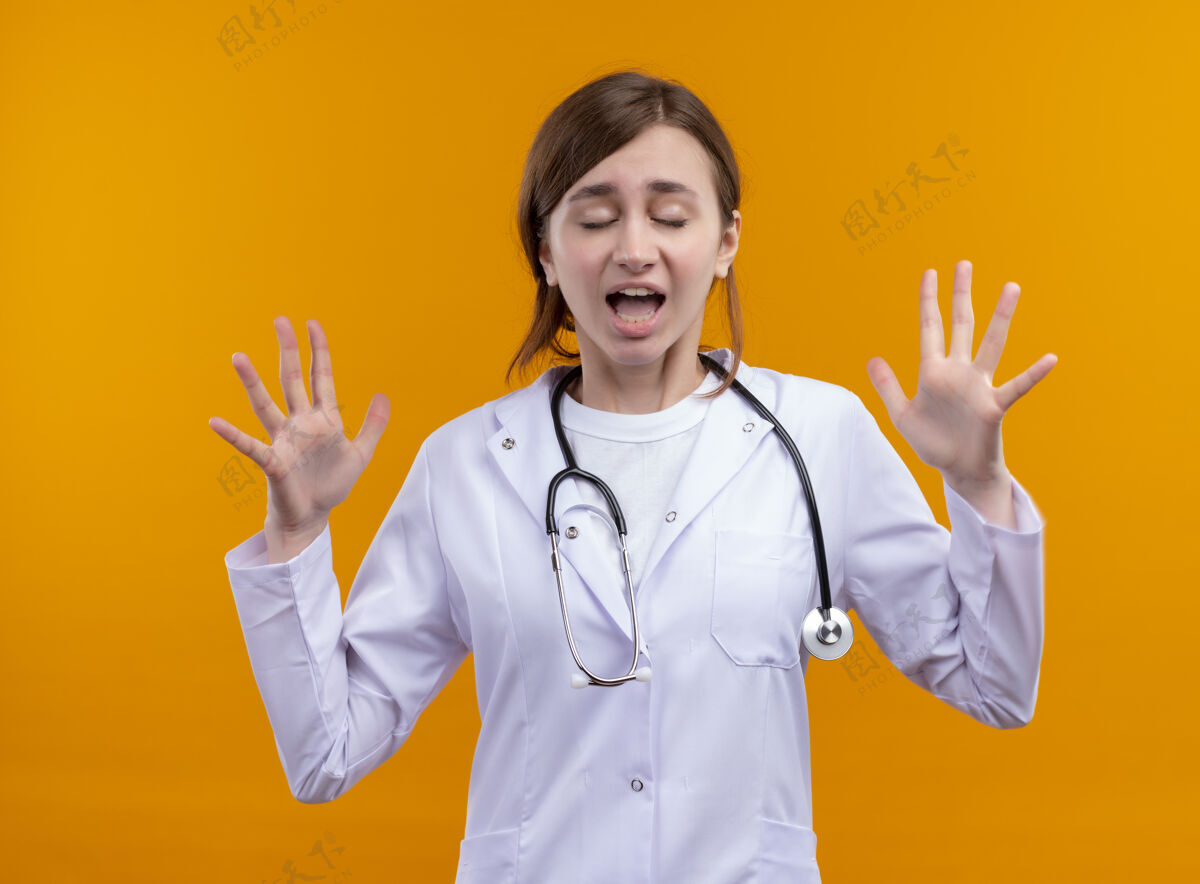 展示打喷嚏的年轻女医生穿着医用长袍和听诊器 在孤立的橙色墙壁上 双手空空 双眼紧闭手医学穿