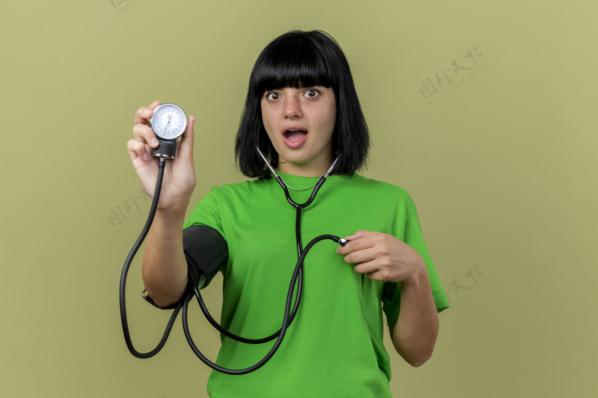 橄榄令人印象深刻的年轻病高加索女孩戴听诊器显示血压计隔离在橄榄绿的墙壁与复制空间印象听诊器血压计