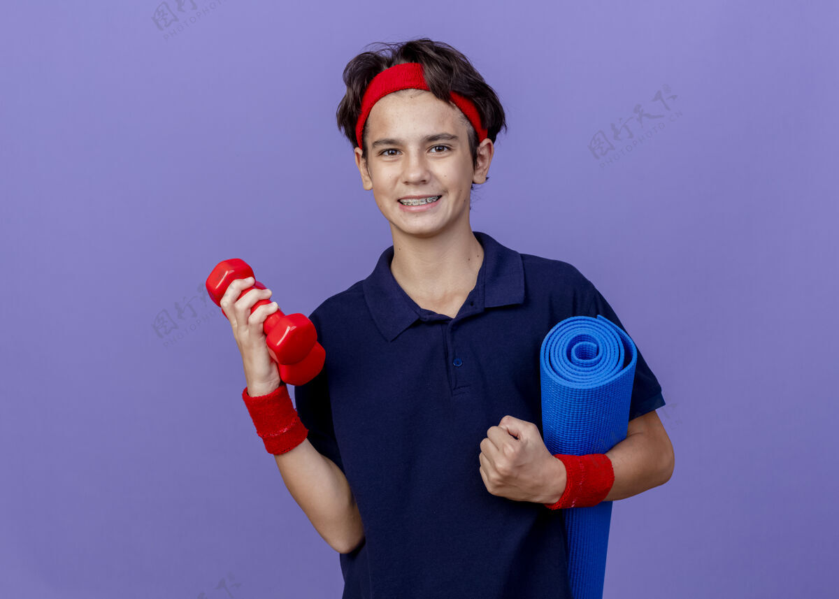 帅气微笑的年轻帅气的运动男孩戴着头带和护腕 戴着牙套 举着哑铃和瑜伽垫 隔离在紫色的墙上 留着复制空间头带垫子运动