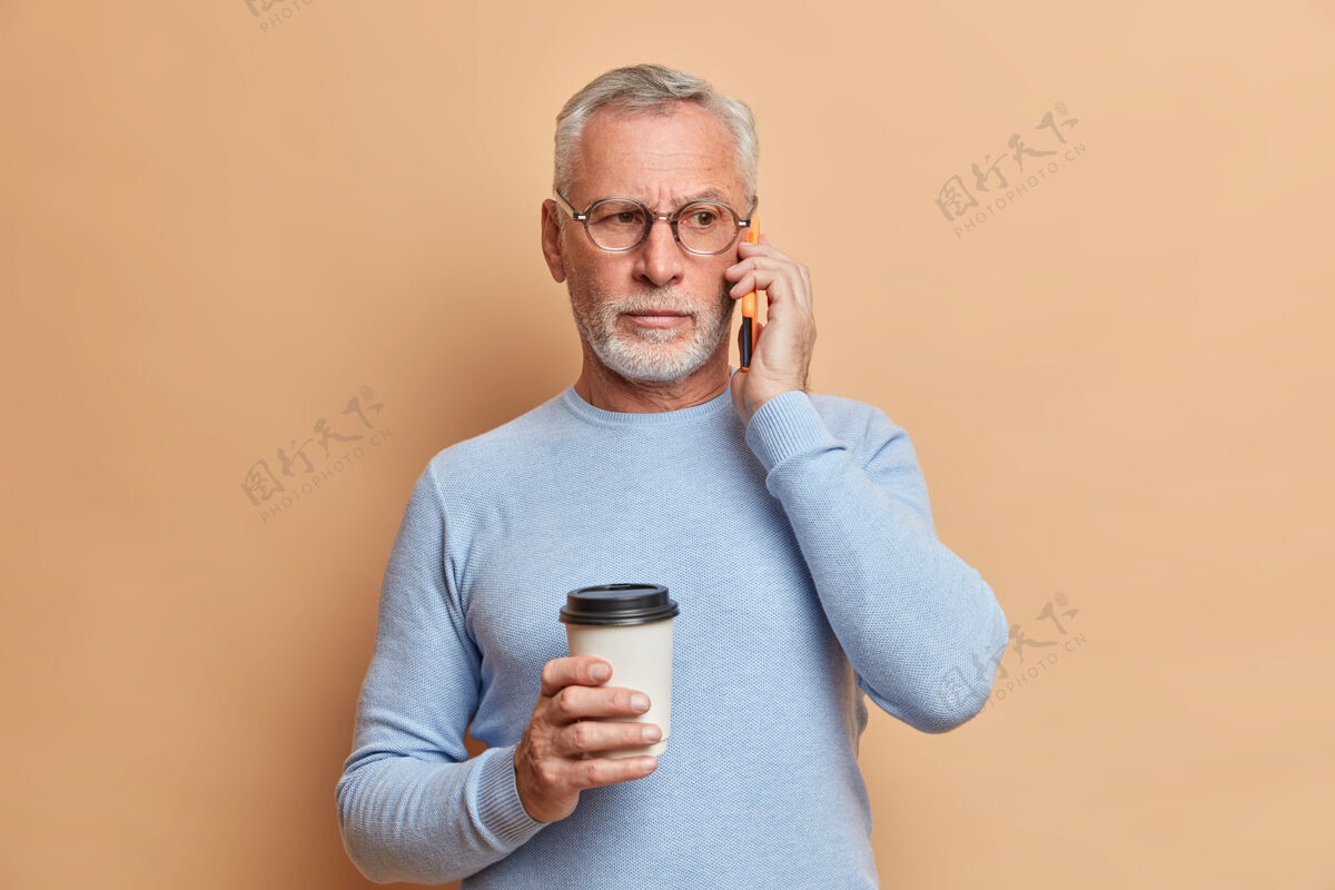 眼镜严肃帅气的大胡子男人打电话聊天 把智能手机放在耳边享受咖啡休息 把目光移开戴着透明眼镜和套头衫隔着棕色的墙成熟电话手机