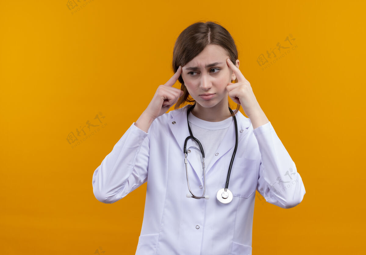 听诊器体贴的年轻女医生穿着医用长袍和听诊器 把手指放在隔离的橙色墙壁上的太阳穴上 并留有复印空间年轻人医生手指