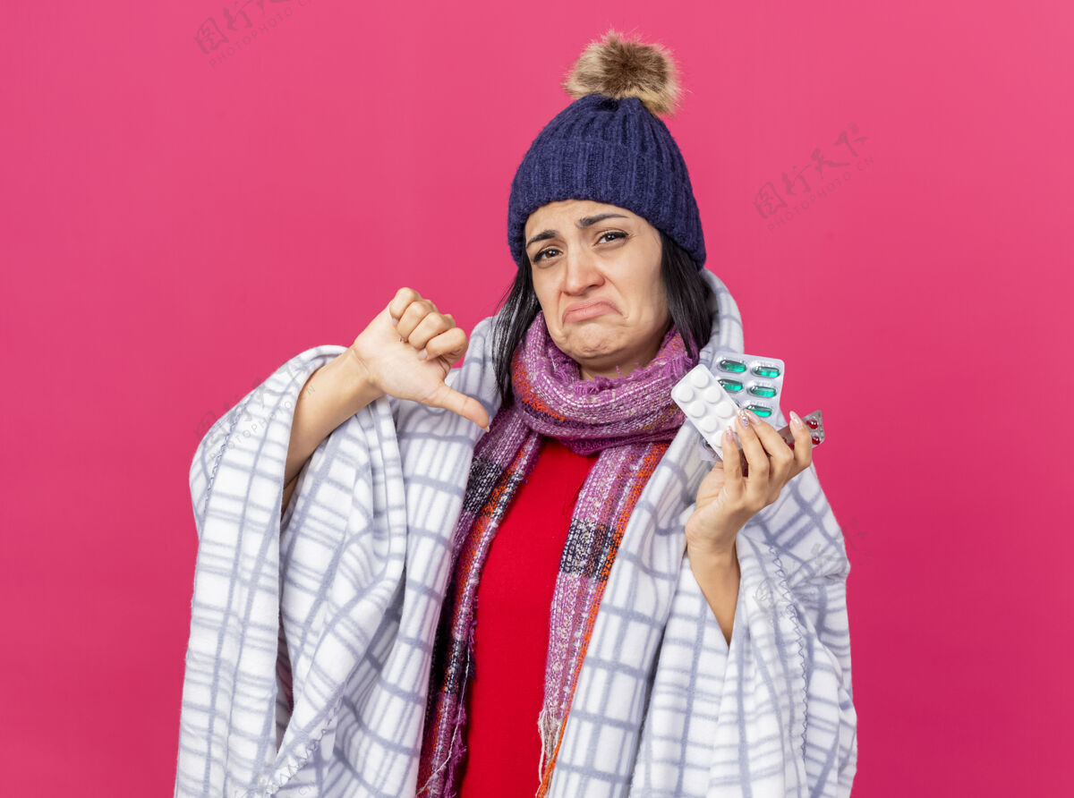 帽子年轻的白种病女孩 戴着冬天的帽子 围着方格布围巾 手里拿着几包药片 拇指朝下 被隔离在深红色的墙上显示深红色药片