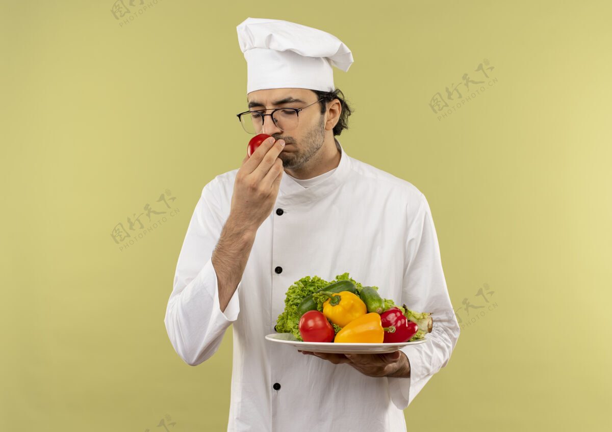 嗅年轻的男厨师闭着眼睛 穿着厨师制服 戴着眼镜 把蔬菜放在盘子里 用手嗅着西红柿 隔离在绿色的墙上 留着复印空间烹饪封闭穿着