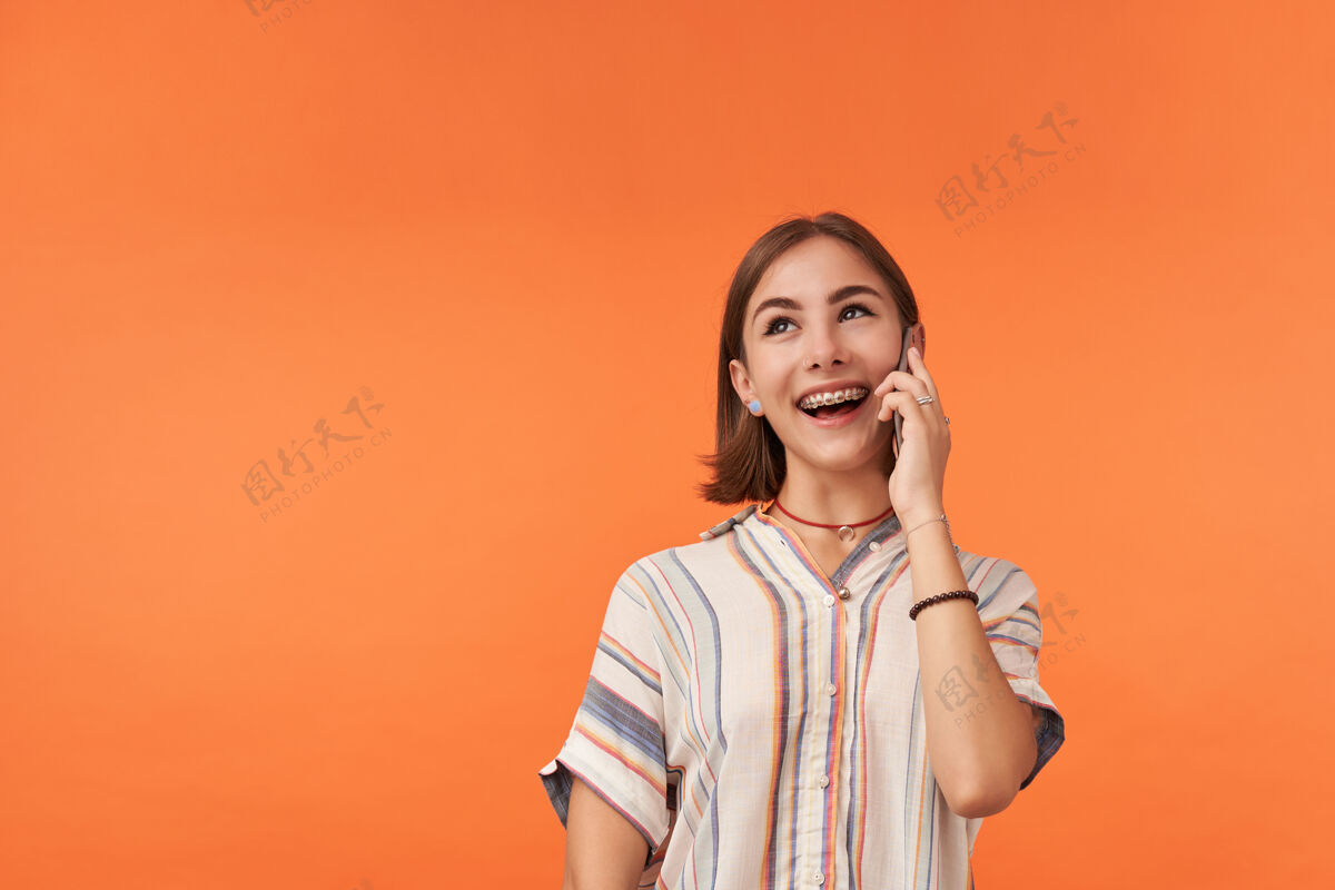 成人年轻女孩在智能手机上思考和交谈穿着条纹衬衫 戴着牙套和手镯站在橙色的墙上微笑着 看着复印区的左上角孤独面部在线