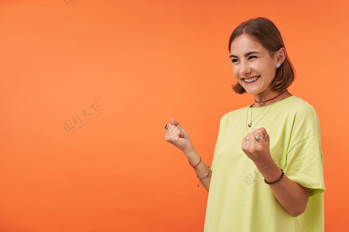 女士女学生 年轻的女士对她所知道的感到兴奋在橘色墙上的复制空间看着左边穿着绿色的t恤 项链 手镯和戒指年轻项链女性