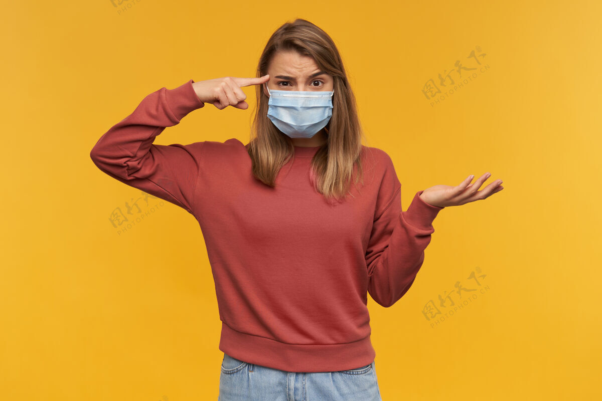 年轻怒气冲冲的年轻女子在医疗保护中用感冒面罩指着她太阳穴上的掌心在掌心上的黄墙提高严格流感