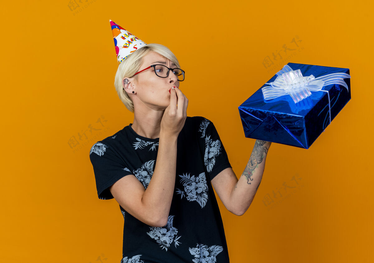 金发年轻的金发派对女孩戴着眼镜 戴着生日帽 拿着礼物盒 做着亲吻的手势 在橙色的背景下与复制空间隔离女孩年轻盒子
