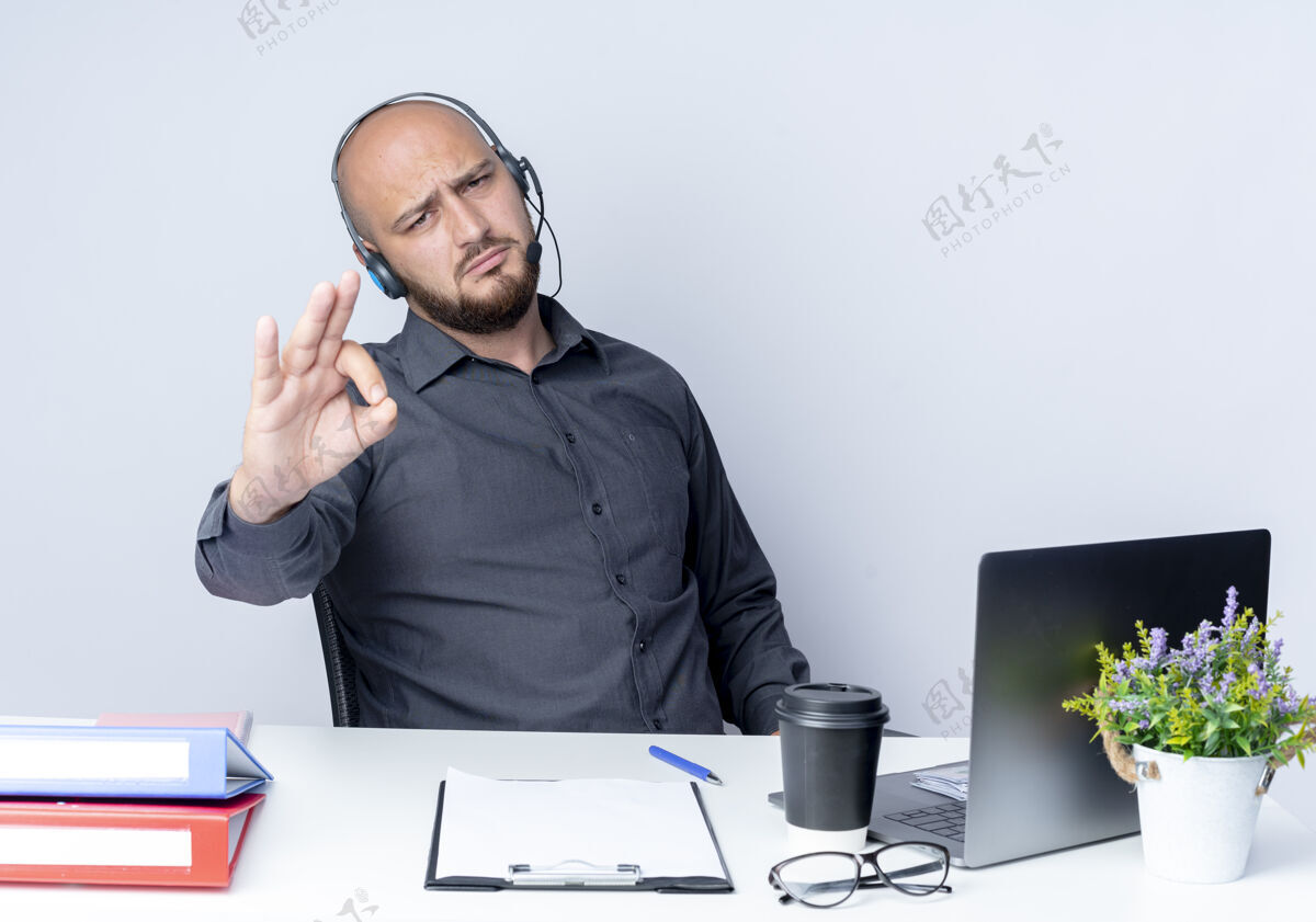 办公桌自信的年轻秃头呼叫中心男子戴着耳机坐在办公桌旁 工作工具做得很好 白色标牌上孤立着中心电话标志