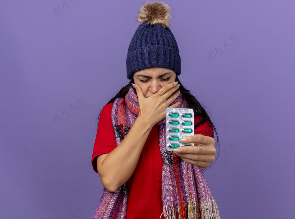 手恼怒的年轻白种人生病的女孩戴着冬天的帽子和围巾 向相机伸出一包胶囊 手放在嘴上 闭着眼睛 隔离在紫色背景上 留有复印空间眼睛疾病保持