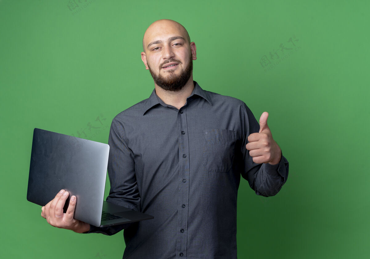 复制自信的年轻秃头呼叫中心男子拿着笔记本电脑和显示拇指孤立在绿色与复制空间中心显示笔记本电脑