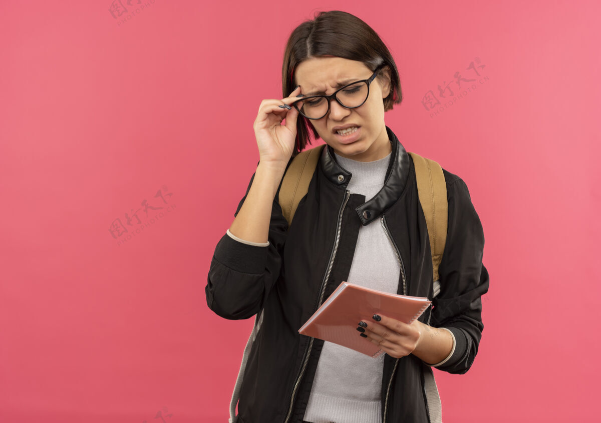 关闭恼怒的年轻女学生戴着眼镜 背着包拿着记事本 她的眼镜闭着眼睛孤立地放在粉红色的复印空间里持有年轻学生
