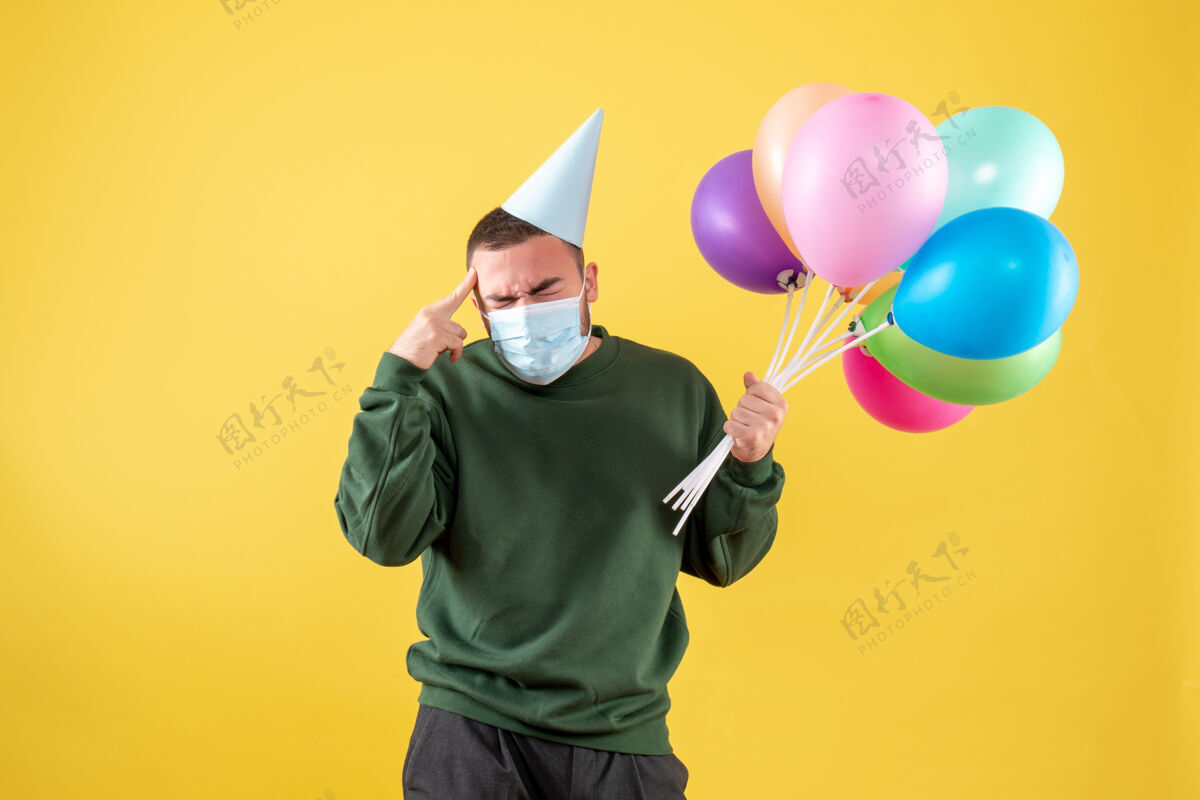 面具正面图：黄色背景上戴着面具手持彩色气球的年轻男性大流行病毒聚会