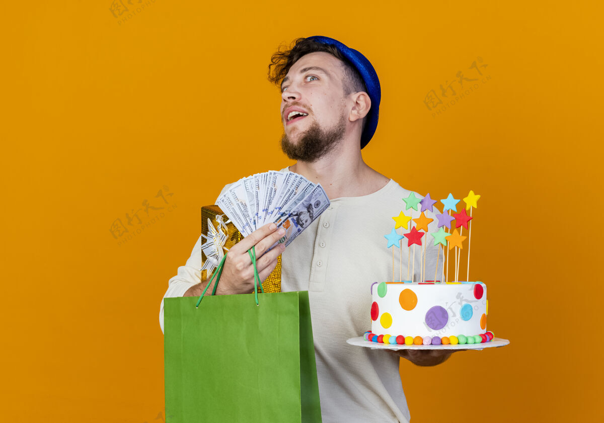 帅哥令人印象深刻的年轻英俊的斯拉夫党家伙戴着党的帽子拿着礼品盒钱纸袋和生日蛋糕与明星看着相机隔离在橙色背景与复制空间包相机派对