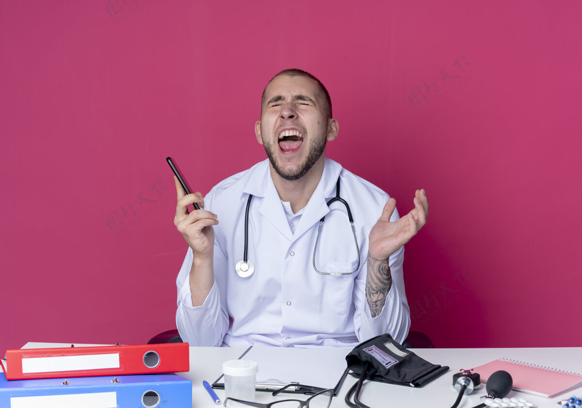 医生愤怒的年轻男医生穿着医用长袍 手持听诊器坐在办公桌旁 手里拿着工作工具 手举着手机 闭着眼睛 与粉色隔离穿坐着空气