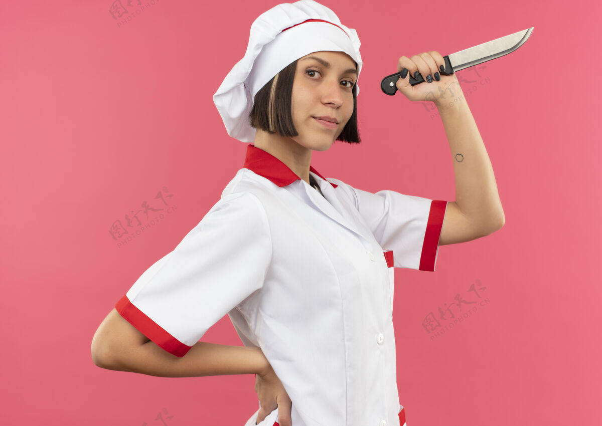 观点自信的年轻女厨师身着厨师制服站在侧视图中 手放在腰上 手持粉红色的刀信心女性腰部