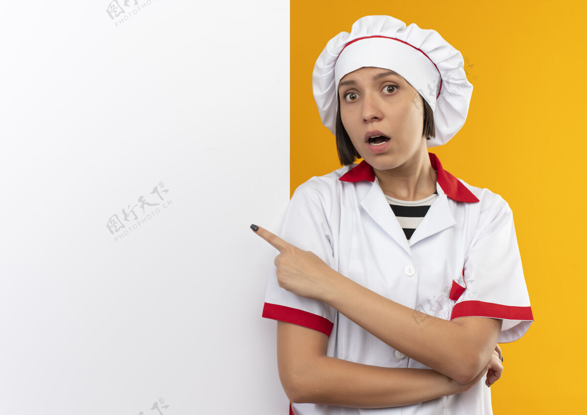 指向穿着厨师制服的年轻女厨师站在白色的墙壁前 指着隔离在橘黄色的墙壁 留着复印空间烹饪复制墙壁